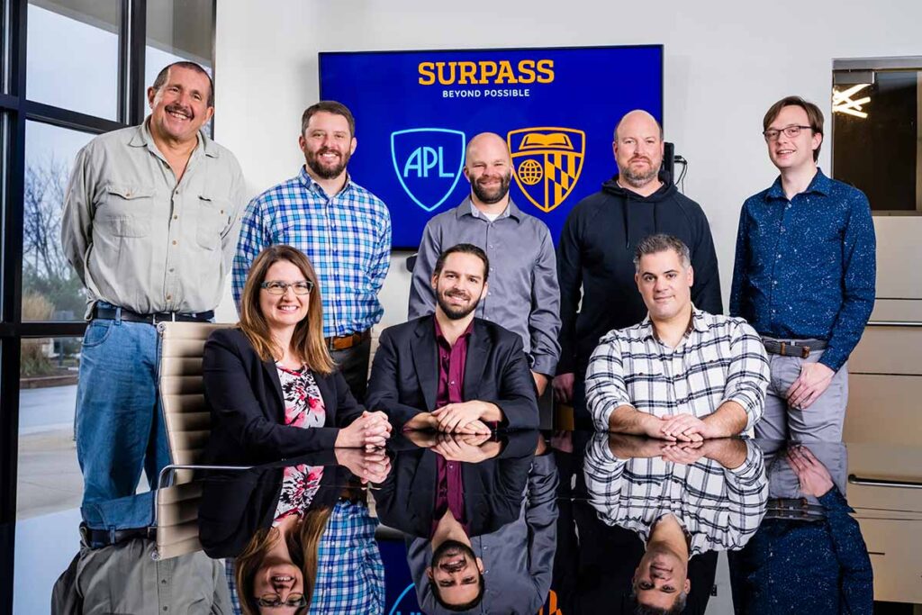 The team working on the CEREBRO project pose together in a conference room. The front row is seated at a reflective black table, and the back row is standing behind them. Team member names are listed in the text below the image.